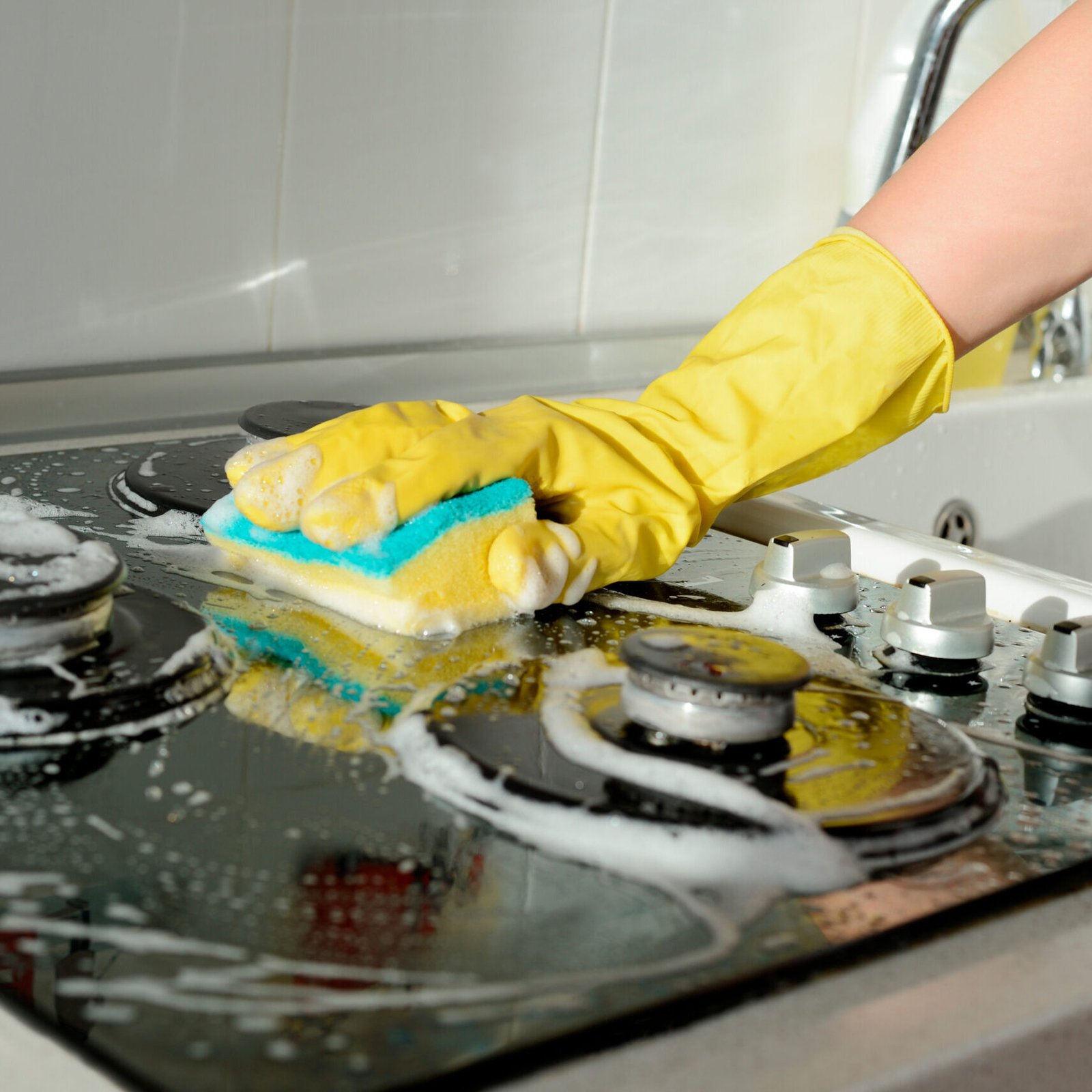 A hand in a yellow rubber glove washes a gas stove on a sunny day. Kitchen cleaning